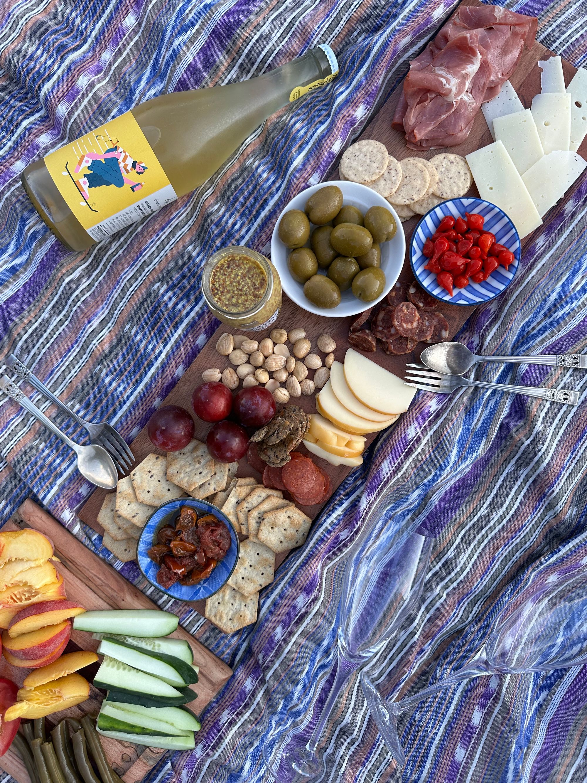 A spread of vegetables, fruits, olives, cheeses, nuts, and natural wines, set upon two cutting boards along a contrasting picnic blanket.