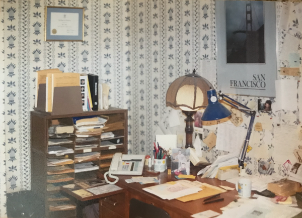 A desk from the '90s sits covered in papers, post-it notes, photos, various lamps, and a phone.