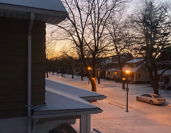 View of the street where a dusting of snow has fallen. Sunrise glows orange on the horizon behind silhouettes of bare trees.